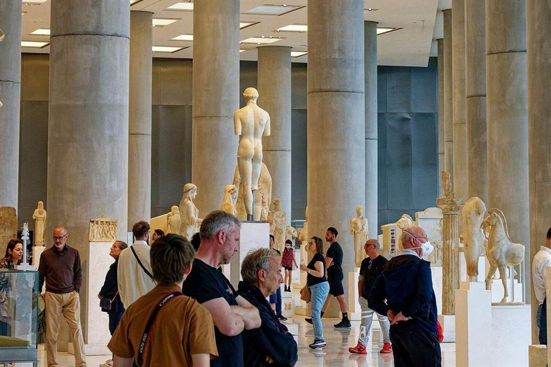 Visitors stand among white marble statues and tall grey concrete columns in a gallery of the Acropolis Museum.