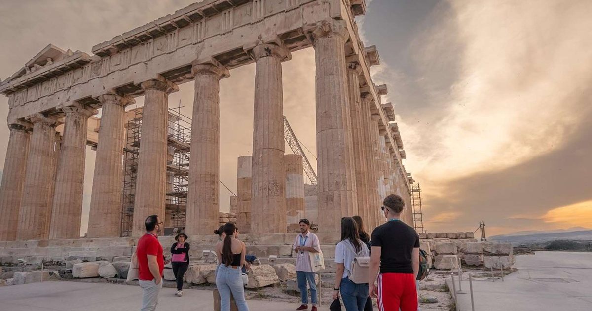 A small group of visitors with a guide stands in front of the Parthenon's Doric columns on the Acropolis of Athens at sunset.