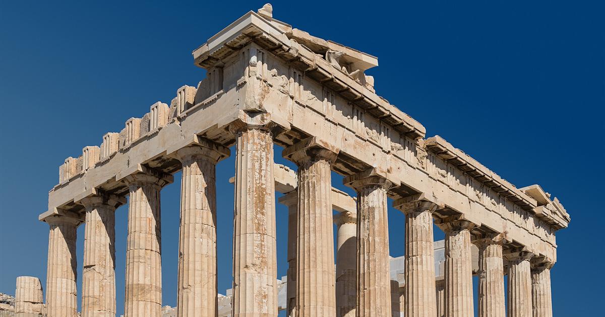 The Parthenon on the Acropolis in Athens, seen at an angle from below, with its Doric columns and carved roof against a deep blue sky.