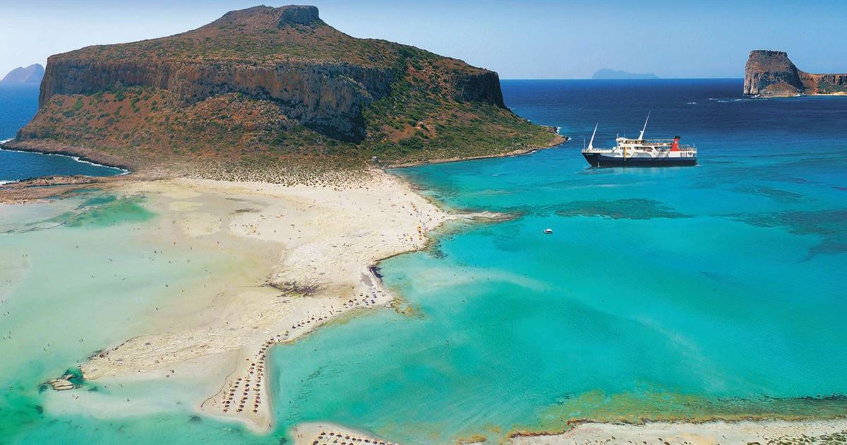 Turquoise water of the Balos lagoon with pale sand and the rocky Gramvousa island with its fortress on top, a tour boat anchored in the distance.