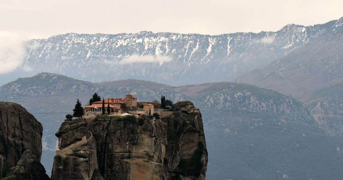 A Meteora monastery with red roofs stands on top of a tall rock pillar, with snow-capped mountains behind it.