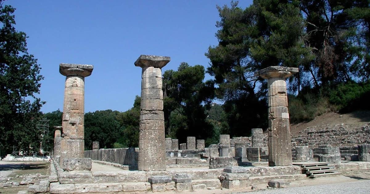 Three tall Doric columns of the Temple of Hera stand on a stone platform under a deep blue sky, with pine trees behind.