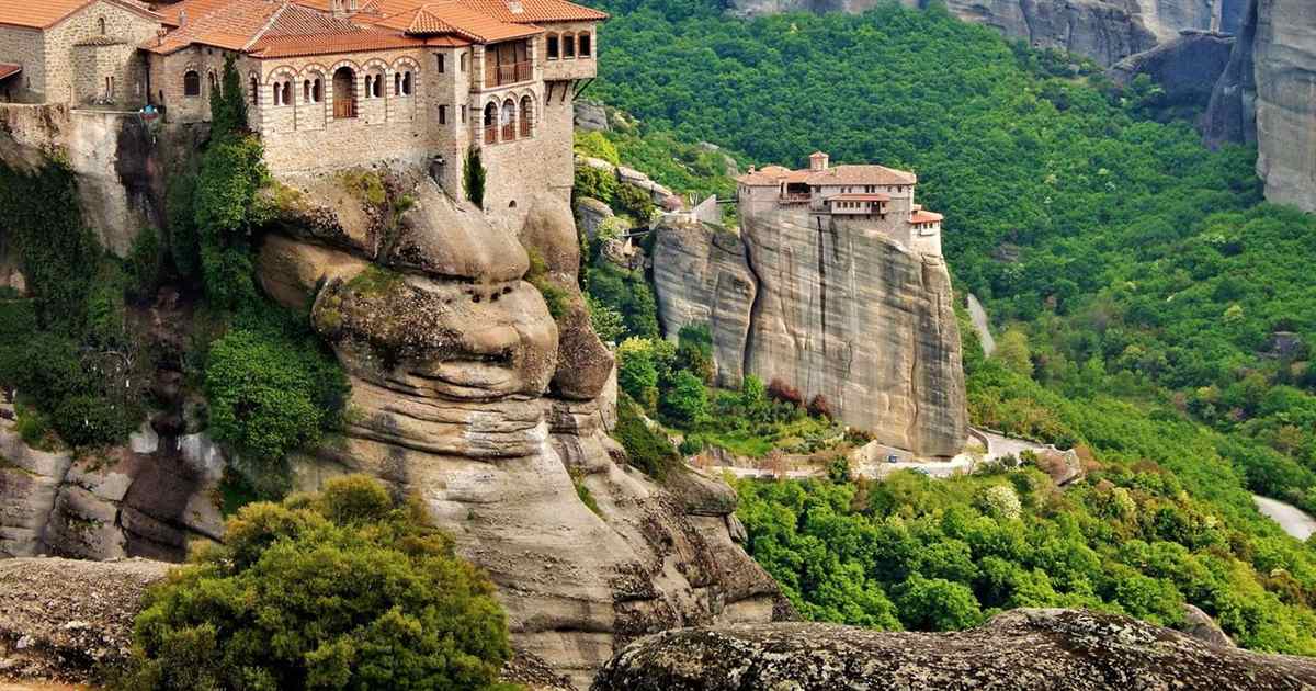 Two monasteries with red tile roofs sit on tall sandstone rock pillars, surrounded by dense green forest, with a deep valley and the Meteora cliffs behind them.