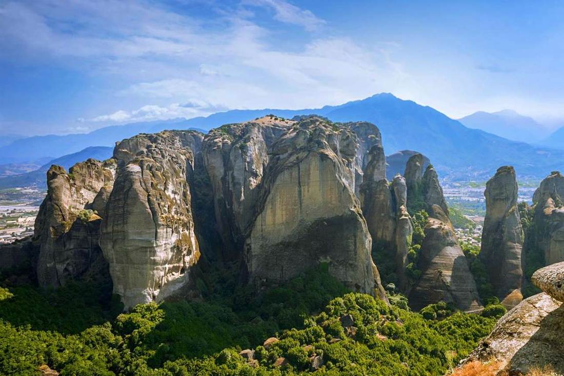 A cluster of tall Meteora rock pillars rising above green forest, with a blue sky and a mountain range behind them.