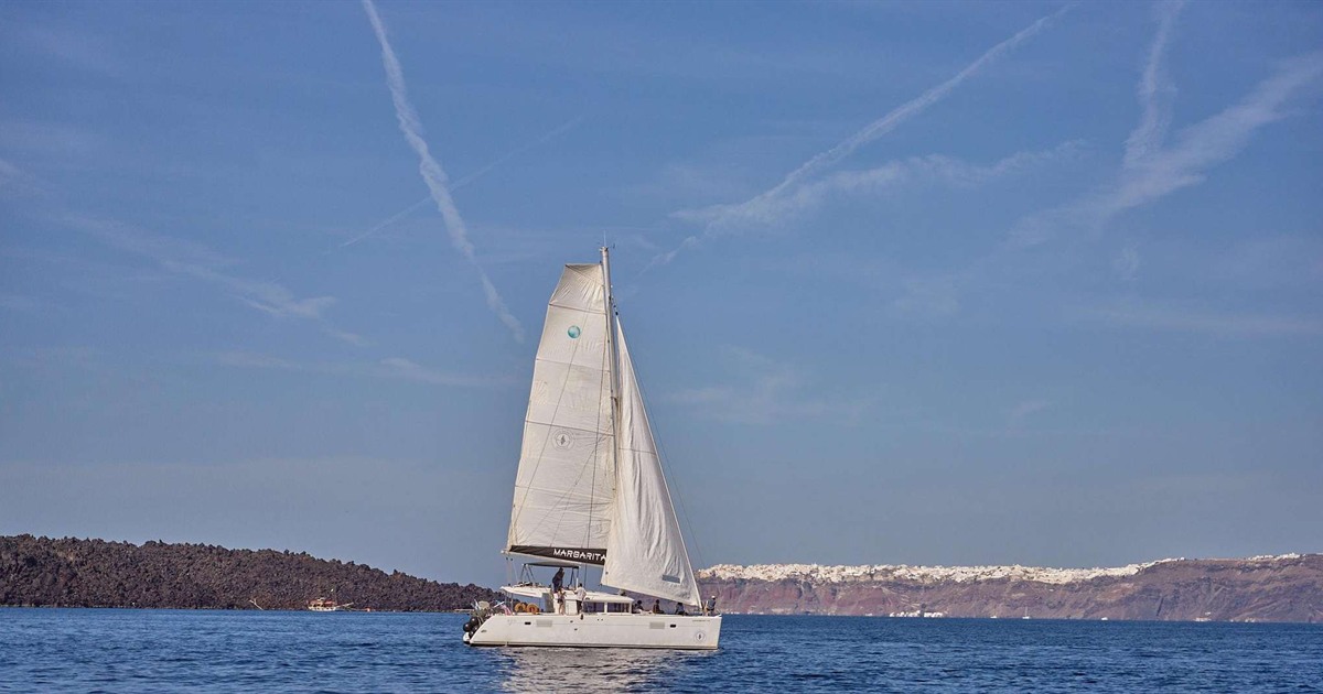 A white two-hulled sailing catamaran with its sails up on calm blue water, with the dark volcanic rock of a small island on the left and the white-topped red cliffs of the Santorini caldera on the right.