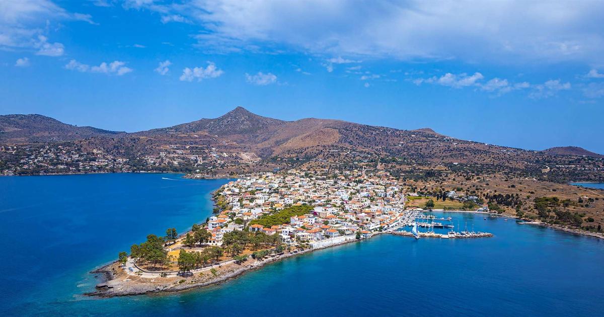 Aerial view of a white-house village on a small peninsula with a marina, deep blue Saronic Gulf water and a brown mountain under a blue sky.