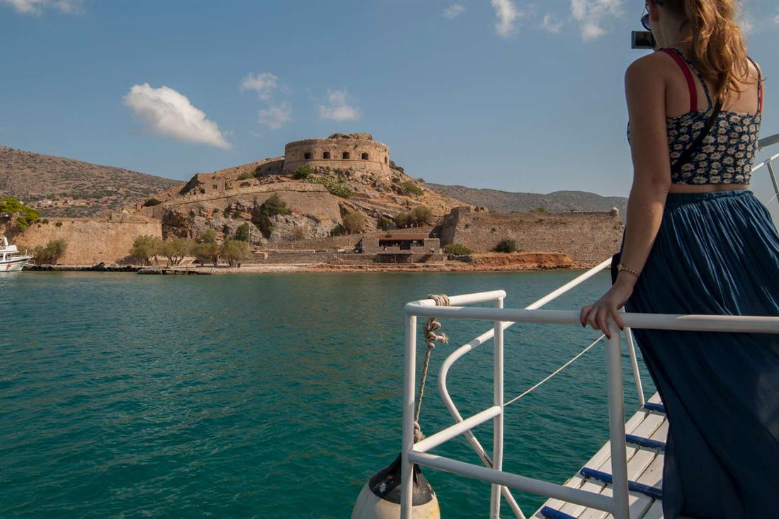 A woman stands at the white railing of a tour boat and looks across the sea at the fortress island of Spinalonga.